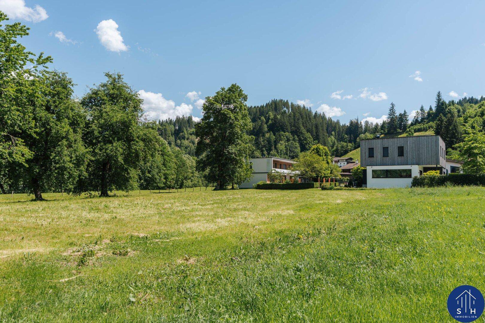 Weitläufige Wiese mit modernen Gebäuden und bewaldeten Hügeln im Hintergrund.