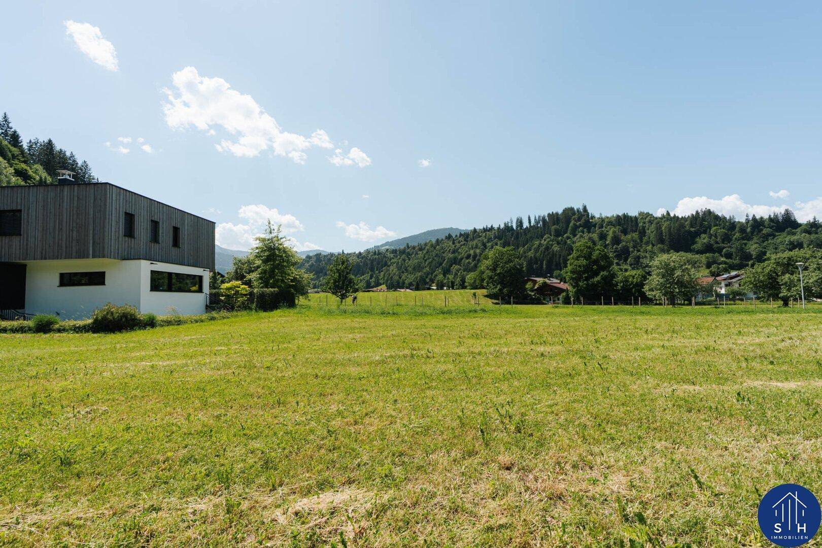 Grüne Wiese vor einem modernen Gebäude mit bewaldeten Bergen unter blauem Himmel.