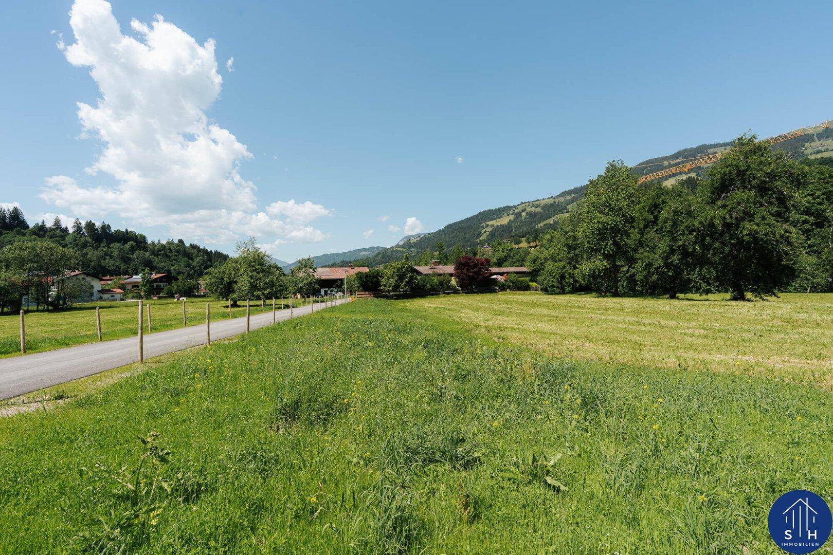 Feldweg entlang einer Wiese mit Blick auf entfernte Häuser und Berge.