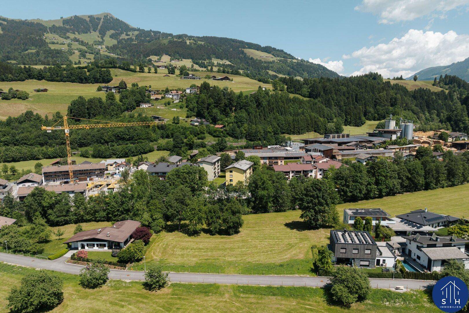 Luftaufnahme eines Dorfes mit Baukränen, umgeben von grünen Wiesen und Wäldern.