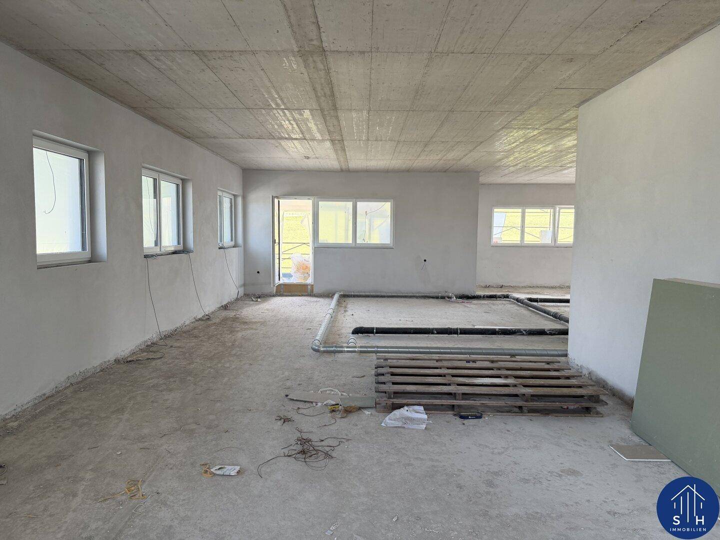 Großer, offener Raum im Rohbau mit vielen Fenstern und Betonboden, bereit für den weiteren Ausbau.