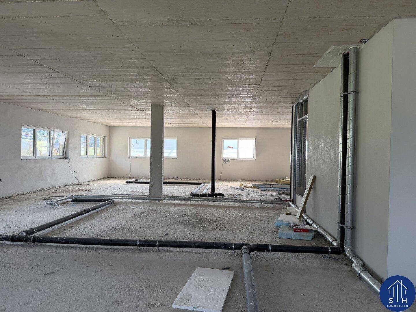 Weiterer Blick in den Rohbau mit sichtbaren Installationen, tragenden Säulen und Fenstern.