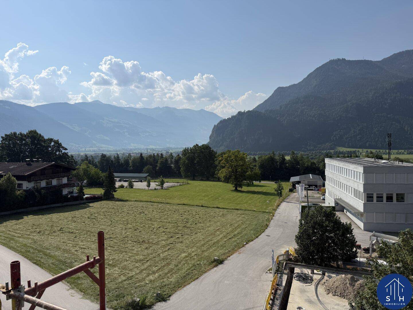 Weitläufiger Blick über grüne Felder und Wiesen mit majestätischen Bergen im Hintergrund unter blauem Himmel.
