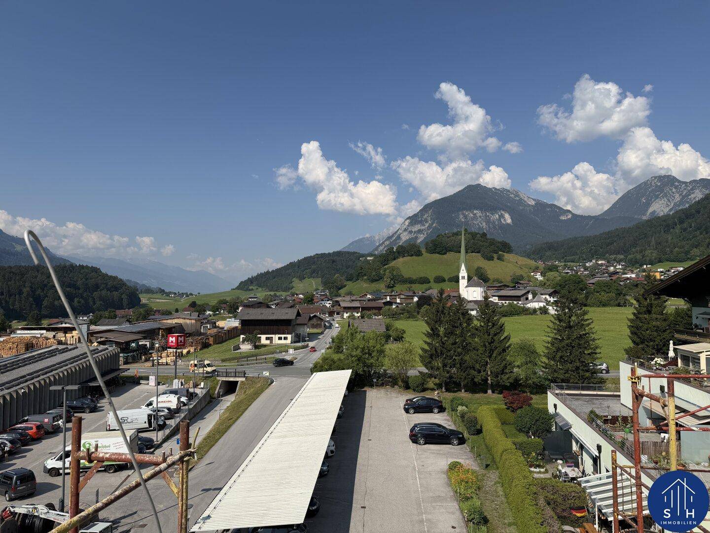 Malerischer Ausblick auf ein Dorf mit Kirche, umgeben von grünen Hügeln und Bergen unter blauem Himmel.