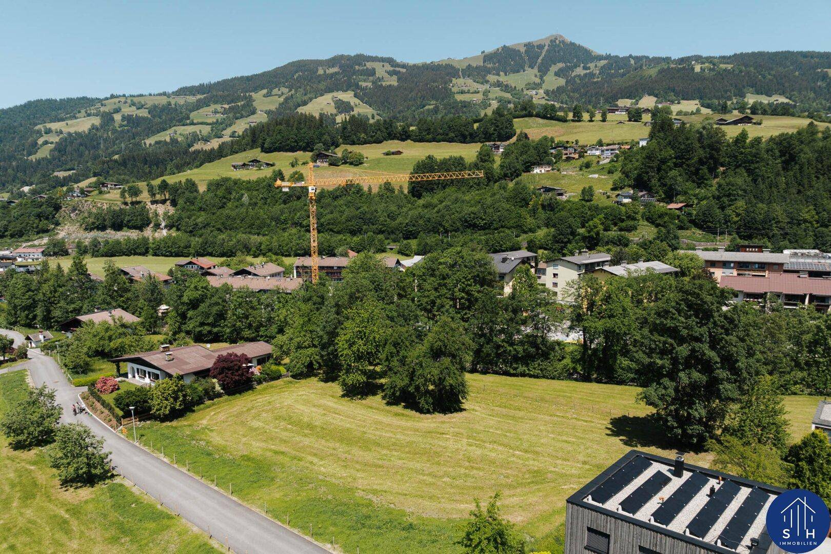 Luftaufnahme einer Wohngegend mit Häusern, grünen Wiesen und Bergen im Hintergrund, inklusive Solaranlage auf einem Dach.