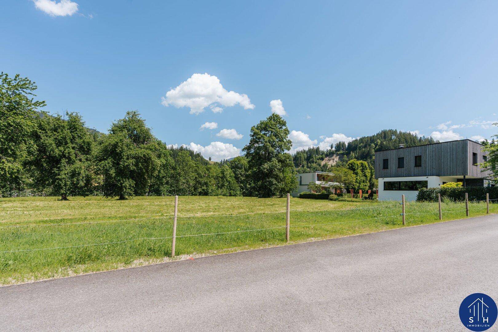 Weitläufige Ansicht eines modernen Hauses inmitten grüner Wiesen mit bewaldeten Hügeln unter klarem Himmel.