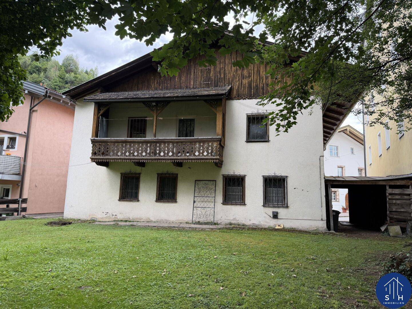 Rückseite des Hauses mit traditionellem Holzbalkon und großem Gartenbereich mit Rasen und Bäumen.