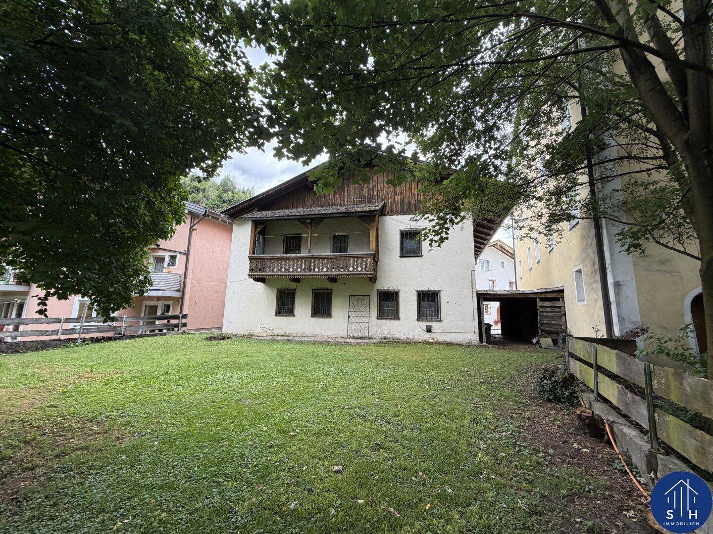 Weitläufiger Garten mit Rasen und altem Baumbestand, angrenzend an die Rückseite des Hauses mit Holzbalkon.