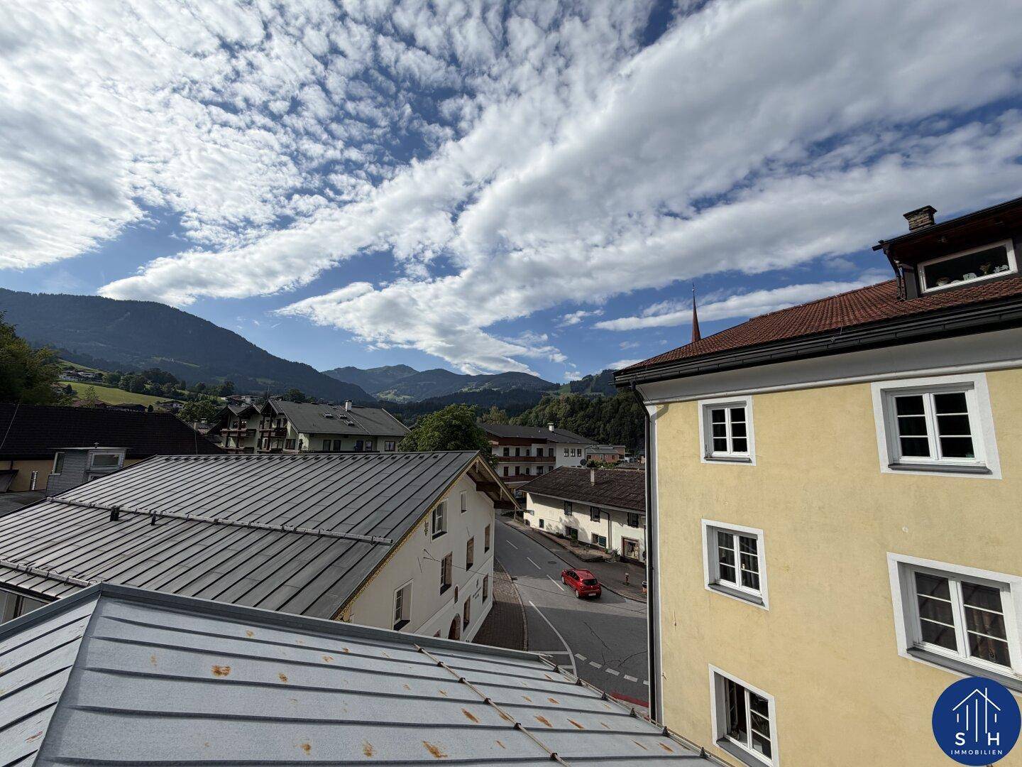 Panoramablick über Dächer und eine Straße mit Bergen im Hintergrund unter bewölktem Himmel.