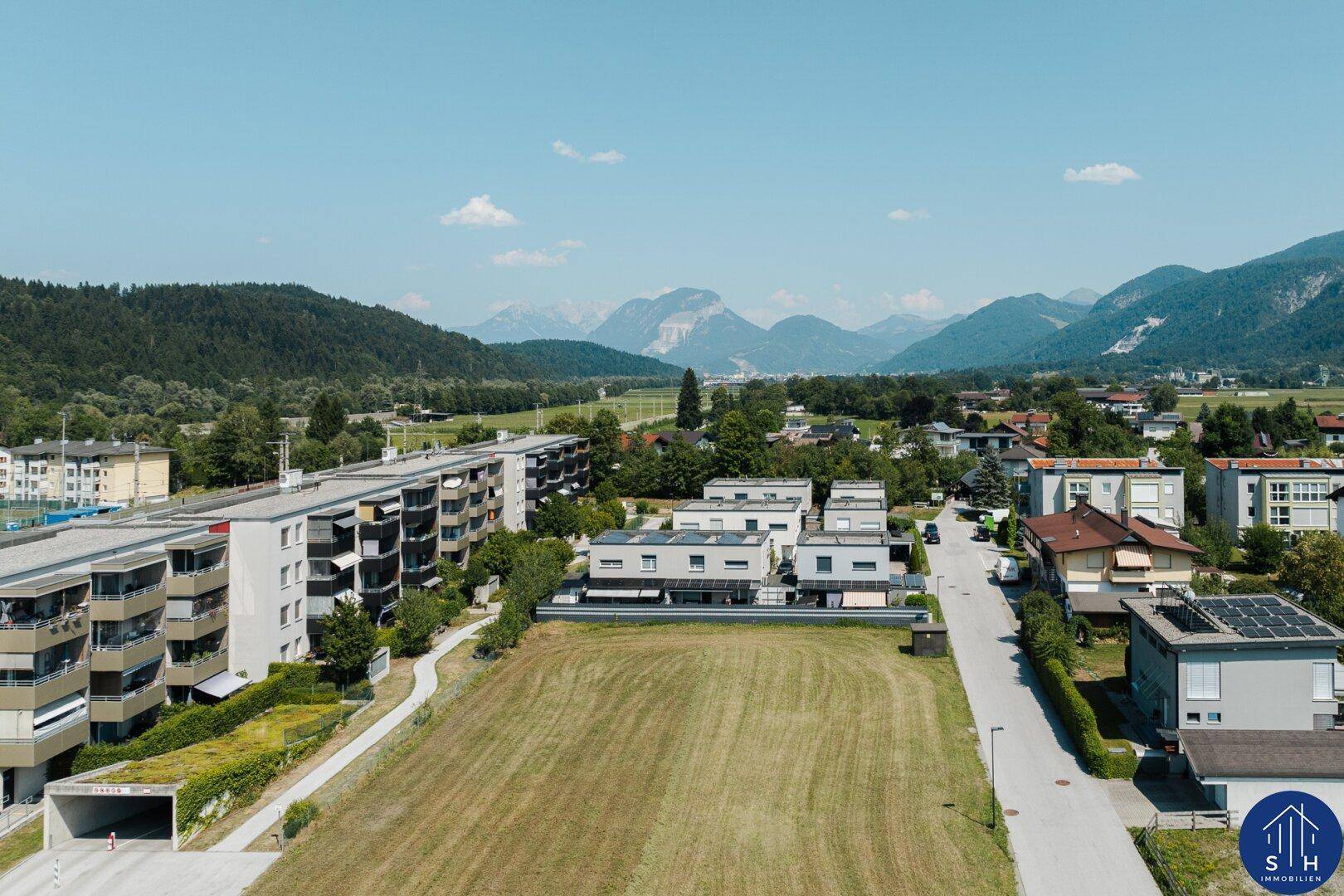 Luftaufnahme einer Wohnsiedlung mit Häusern, Feldern und Bergen im Hintergrund.