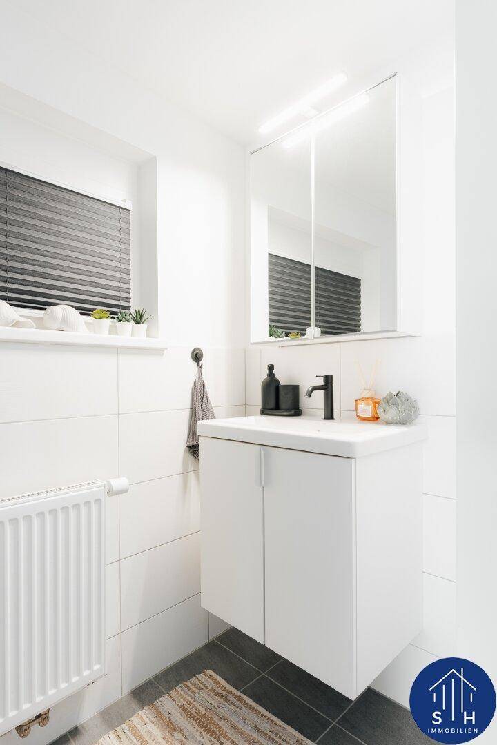 Kleines Badezimmer mit Waschtisch, Spiegelschrank und Fenster, in Weiß und Grau gefliest.