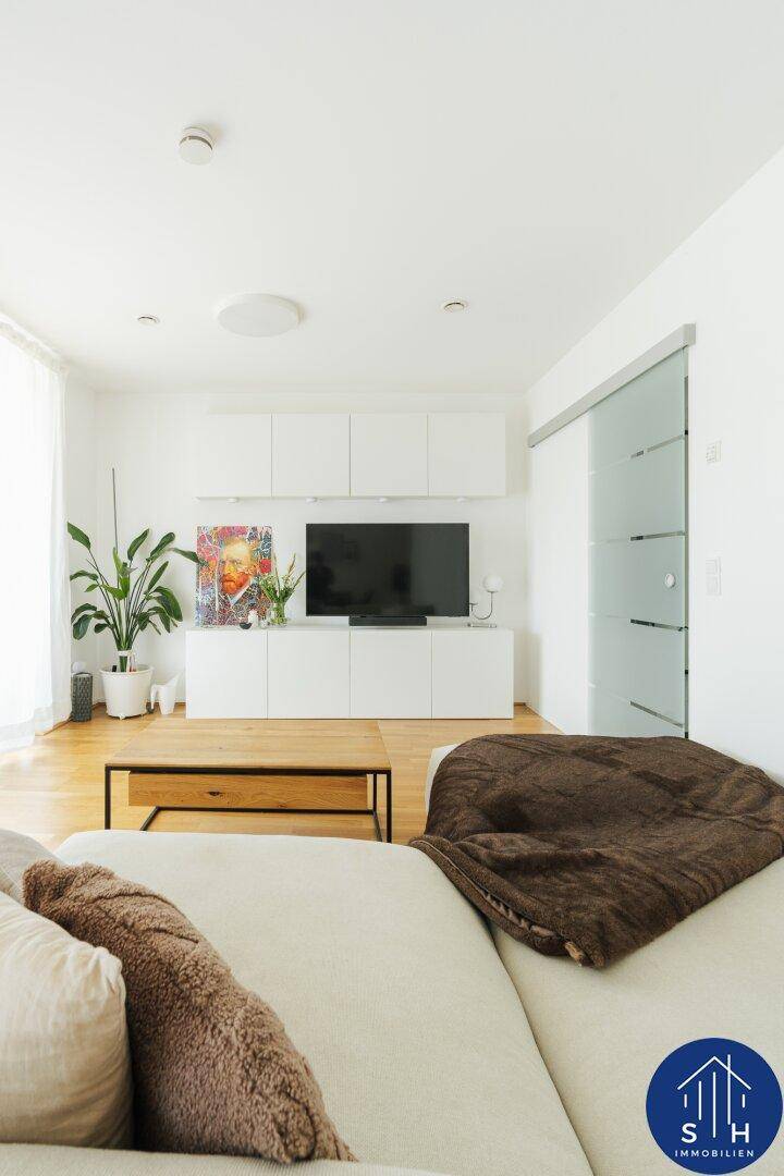 Helles Wohnzimmer mit TV-Möbeln, Zimmerpflanze und Holztisch, durch Glasschiebetür verbunden.