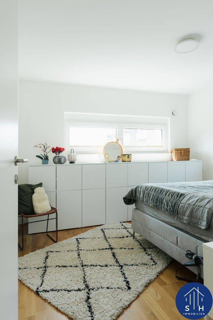 Helles Schlafzimmer mit Bett, integrierten weißen Schränken und einem großen Fenster.