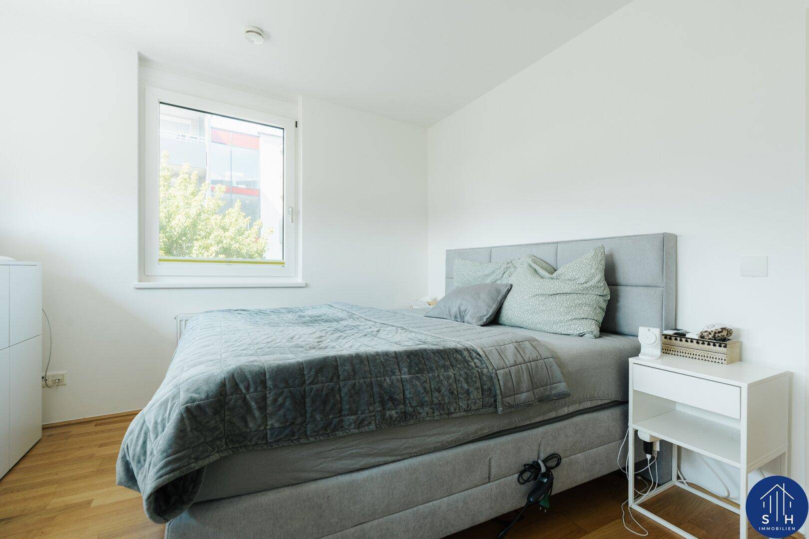 Ruhiges Schlafzimmer mit Bett, Nachttisch und einem Fenster, das Tageslicht hereinlässt.