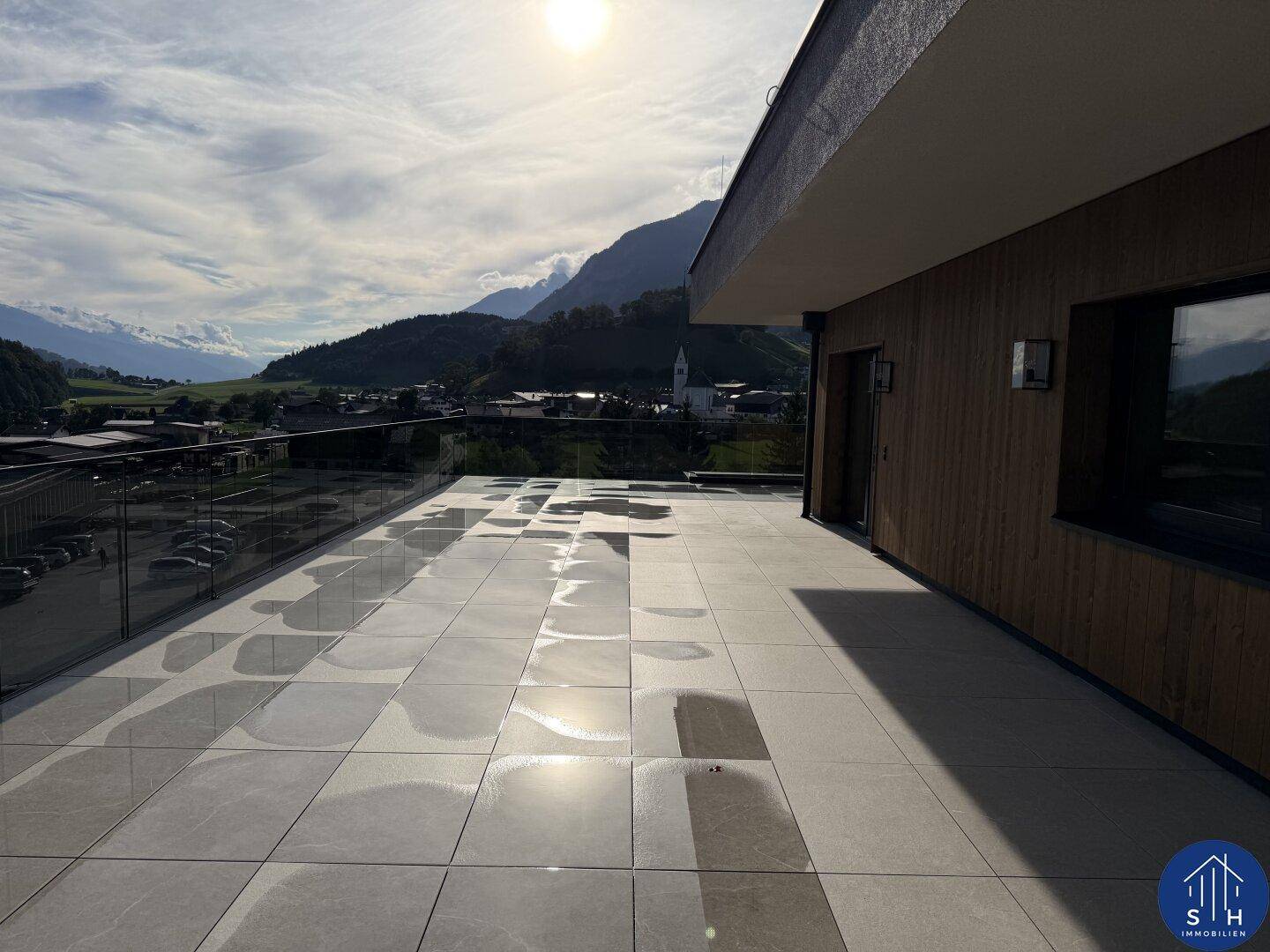 Sonnige Terrasse mit Fliesenboden und Glasgeländer, bietet einen weiten Blick auf die umliegende Landschaft und Berge.