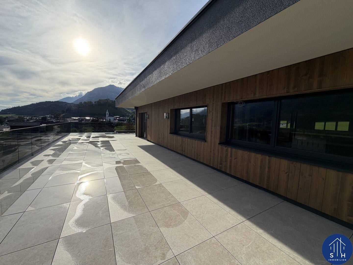 Sonnige Terrasse mit moderner Holzfassade und Fliesenboden, umgeben von einer beeindruckenden Berglandschaft.