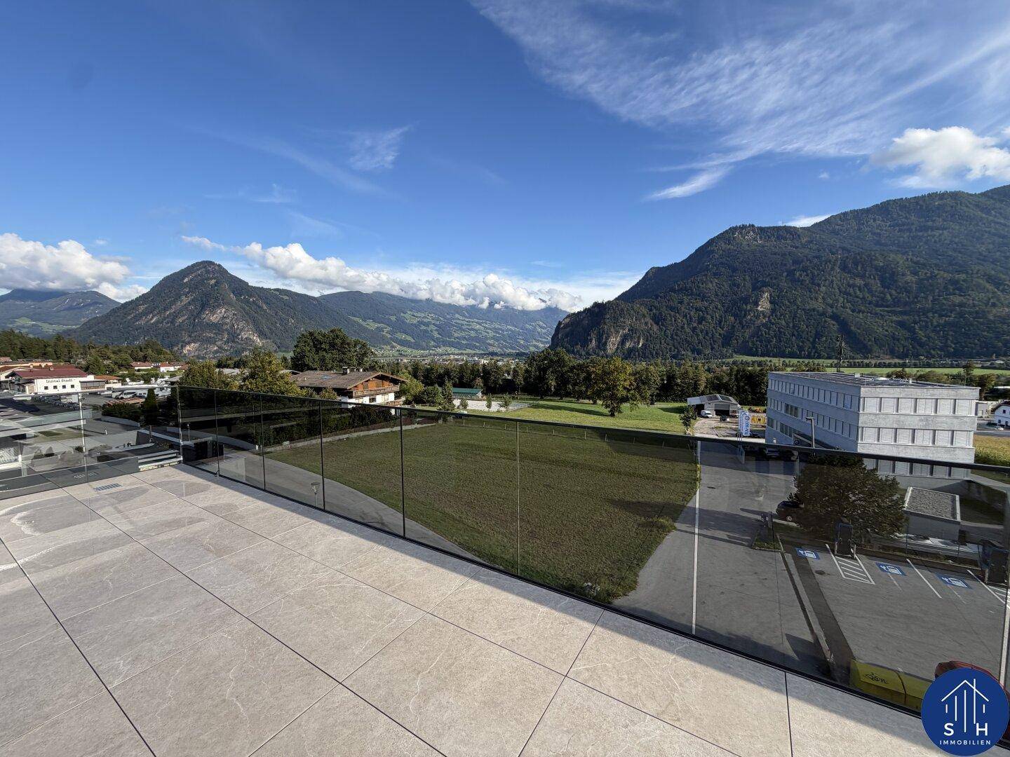 Geräumige Terrasse mit Fliesenboden und Glasgeländer, bietet einen weiten Blick auf grüne Felder und Berge.