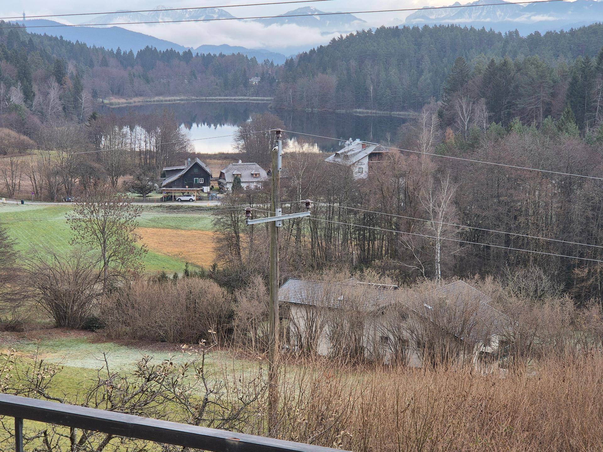 Nahaufnahme des Sees mit umliegenden Häusern und bewaldeten Ufern, unter einem grauen Himmel.