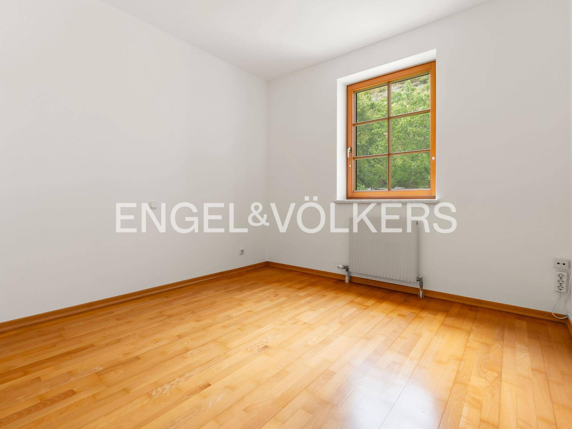 Ruhiges Zimmer mit Parkettboden, weiß gestrichenen Wänden und einem Fenster mit Blick ins Grüne.