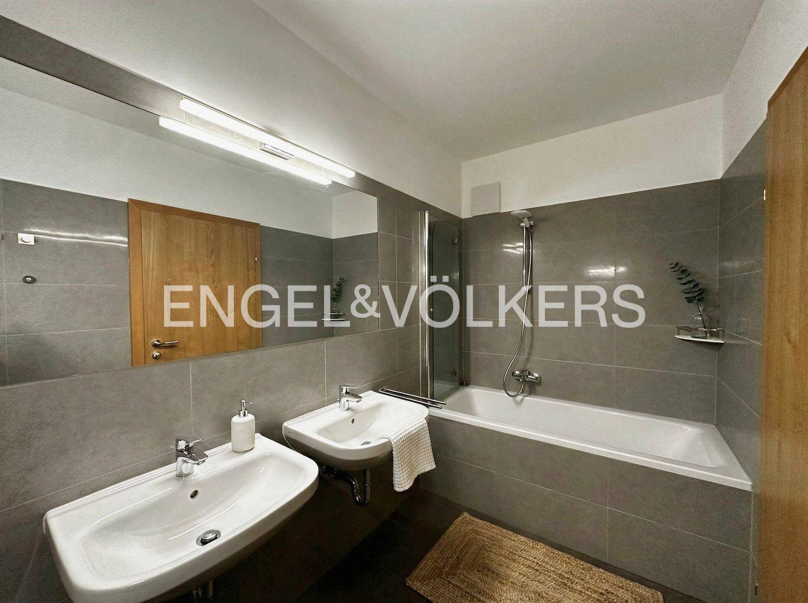 Modernes Badezimmer mit zwei Waschbecken, Badewanne mit Duschfunktion und grauen Fliesen.