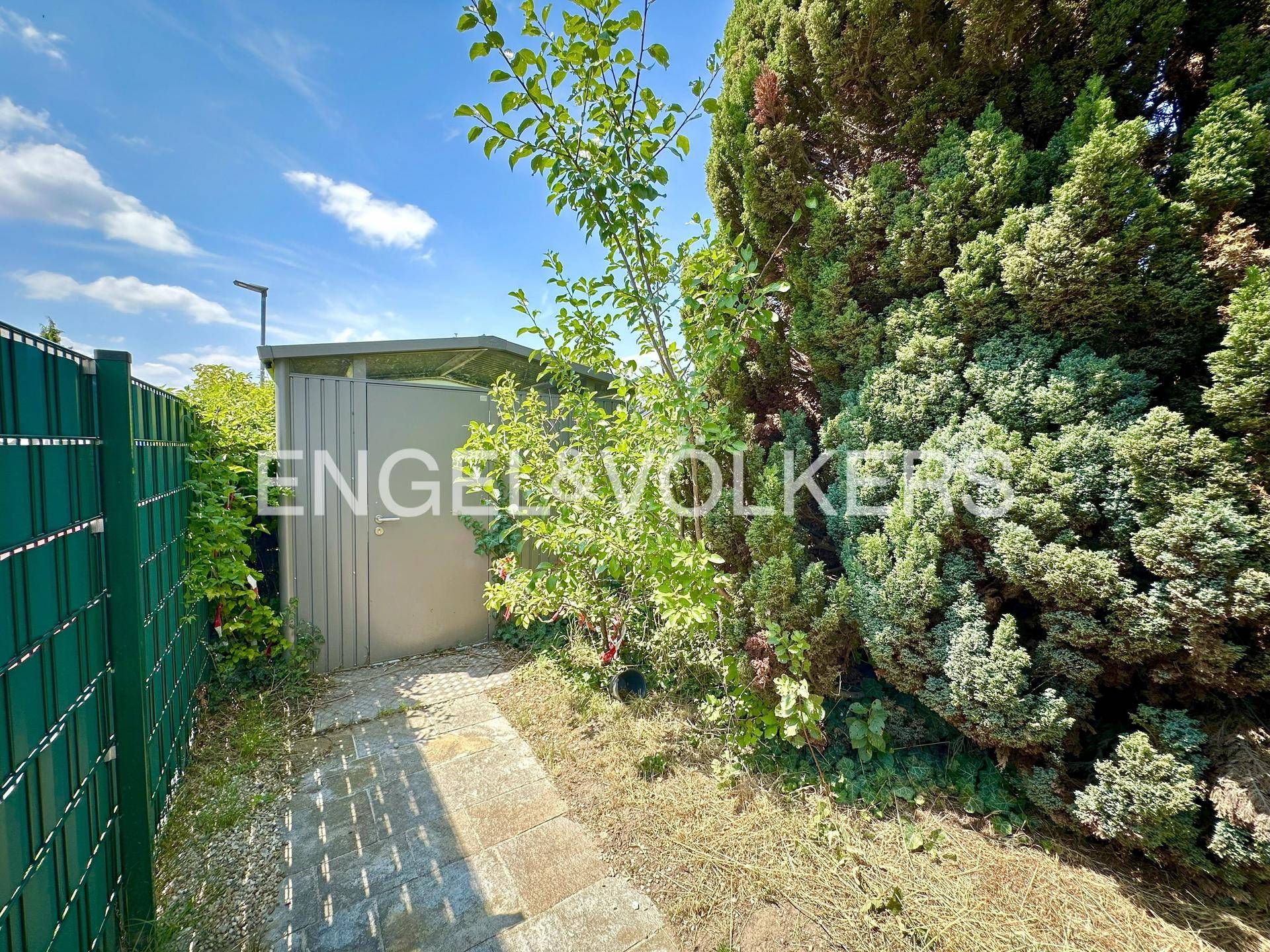 Kleiner Gartenschuppen neben einer Hecke und einem Steinweg in einem sonnigen Gartenbereich.