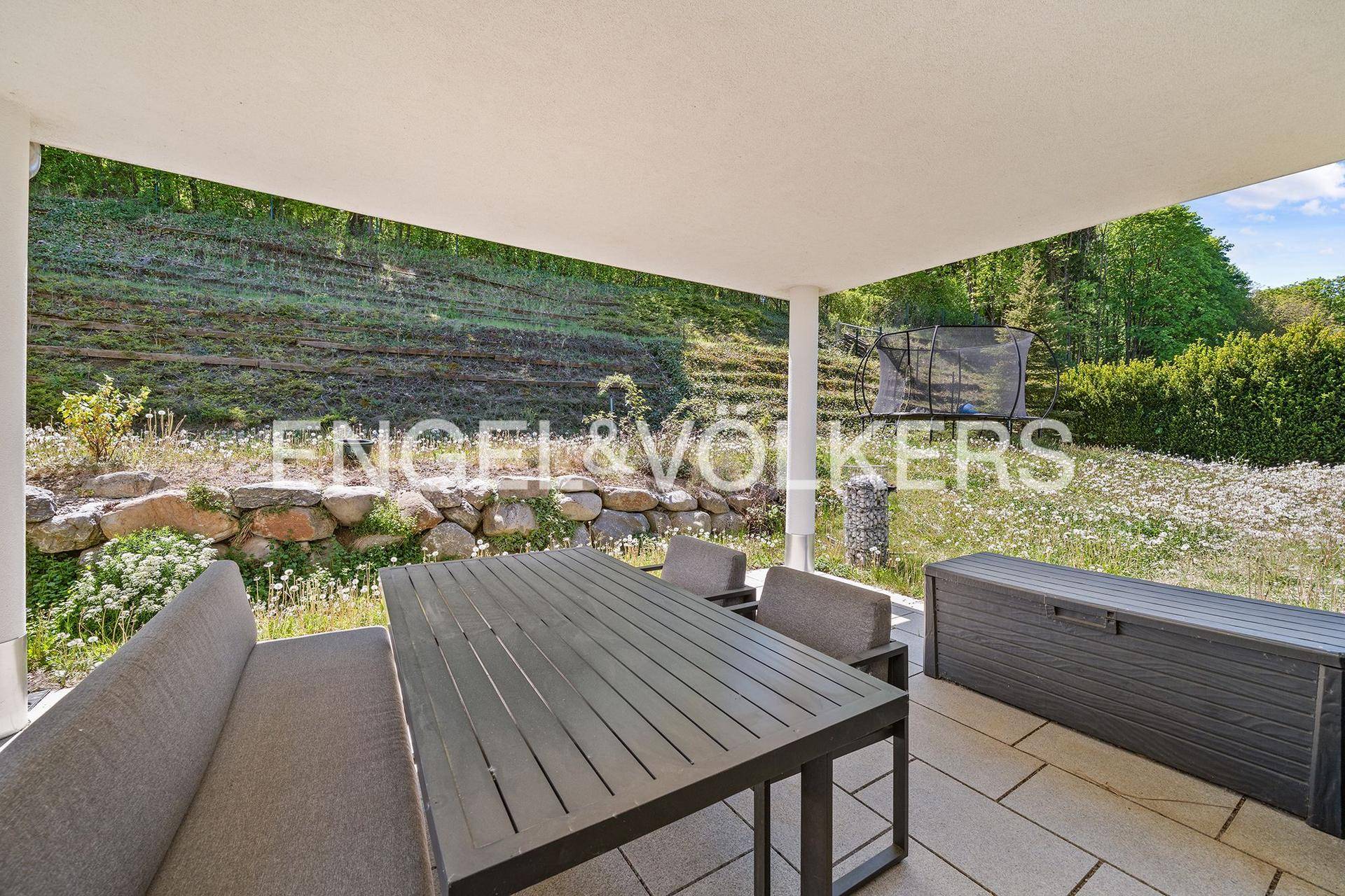 Überdachte Terrasse mit Esstisch und Sitzgelegenheiten, umgeben von einer grünen Hanglandschaft.