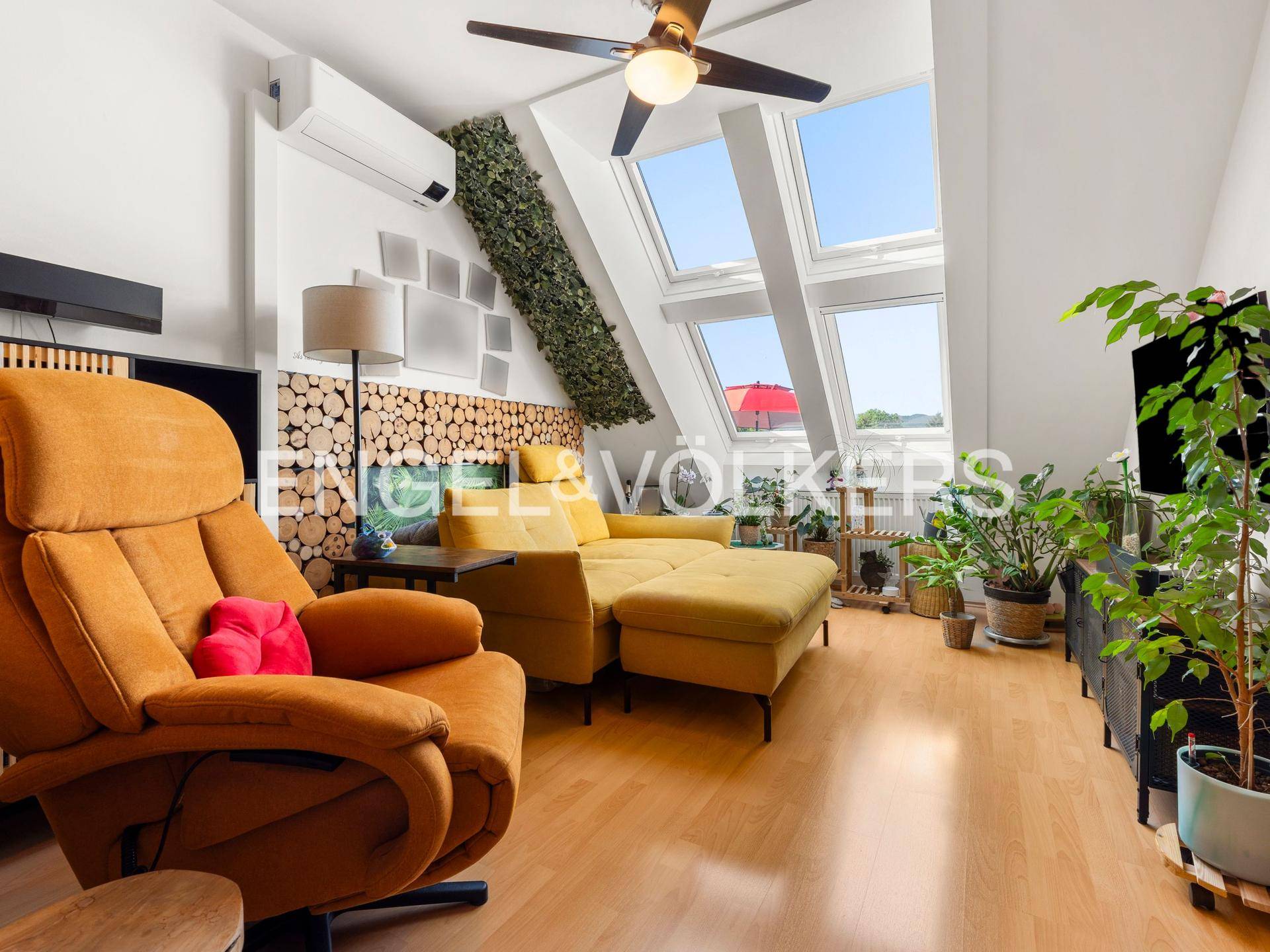 Gemütliches Wohnzimmer im Dachgeschoss mit großen Dachfenstern, gelbem Sofa und Deckenventilator.