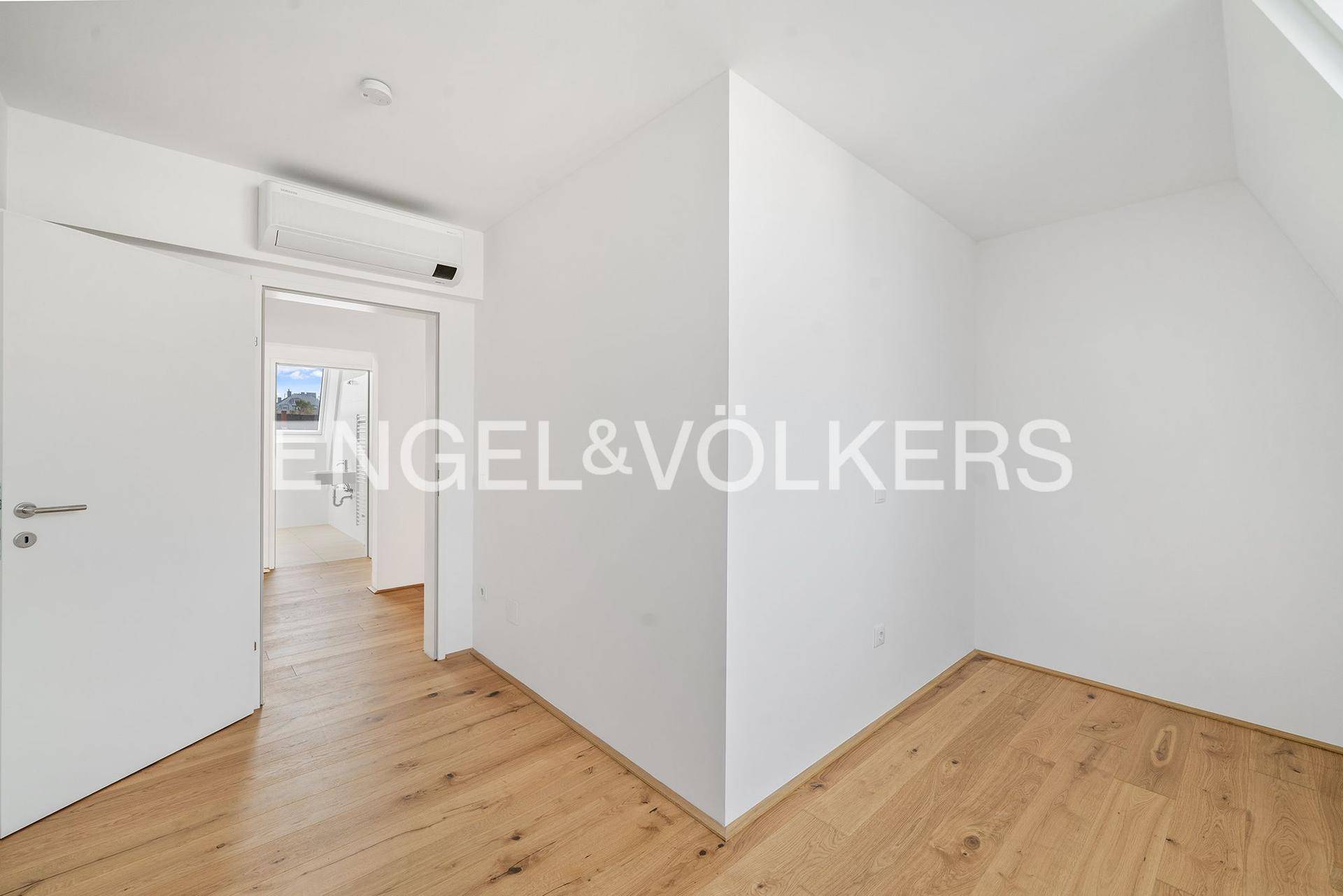 Helles, leeres Zimmer mit Holzboden und Klimaanlage, mit Blick in den Flur.