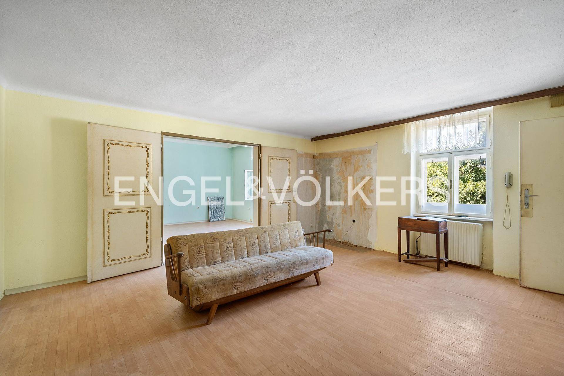 Geräumiges Wohnzimmer mit Parkettboden, einem Sofa und Blick ins Grüne, renovierungsbedürftig.