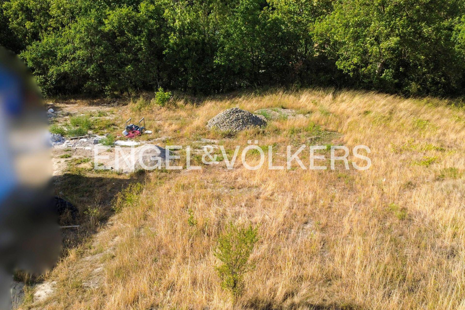 Trockenes Wiesengrundstück mit einem Kieshaufen und Baustellenmaterial im Vordergrund.