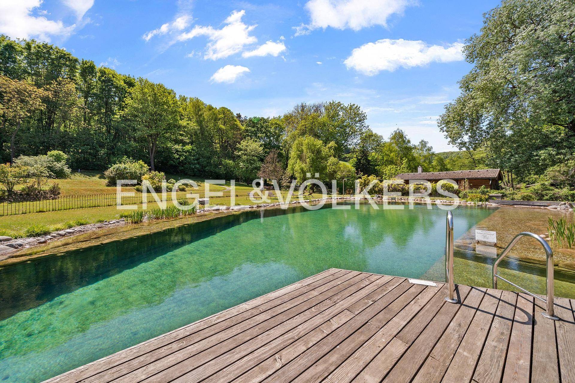 Großer Naturpool mit Holzdeck und üppiger grüner Umgebung unter blauem Himmel.