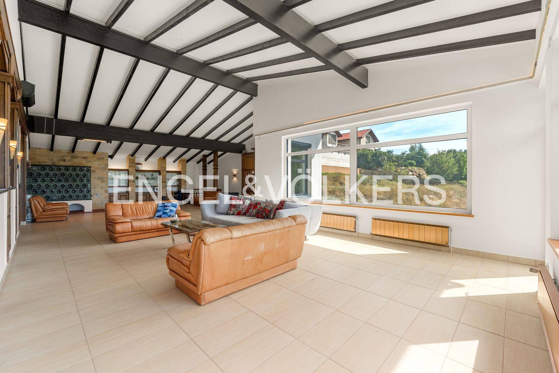 Helles Wohnzimmer mit Fliesenboden, sichtbaren Deckenbalken und großen Fenstern zur Natur.