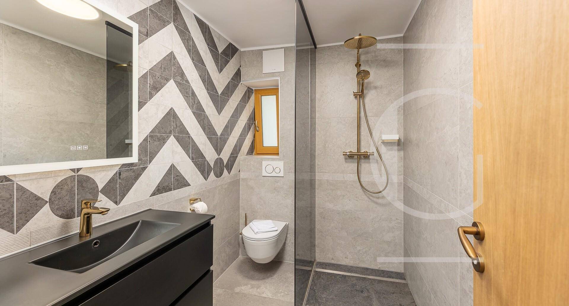 Modernes Badezimmer mit begehbarer Dusche, geometrischen Fliesen und stilvollen Armaturen in Gold.
