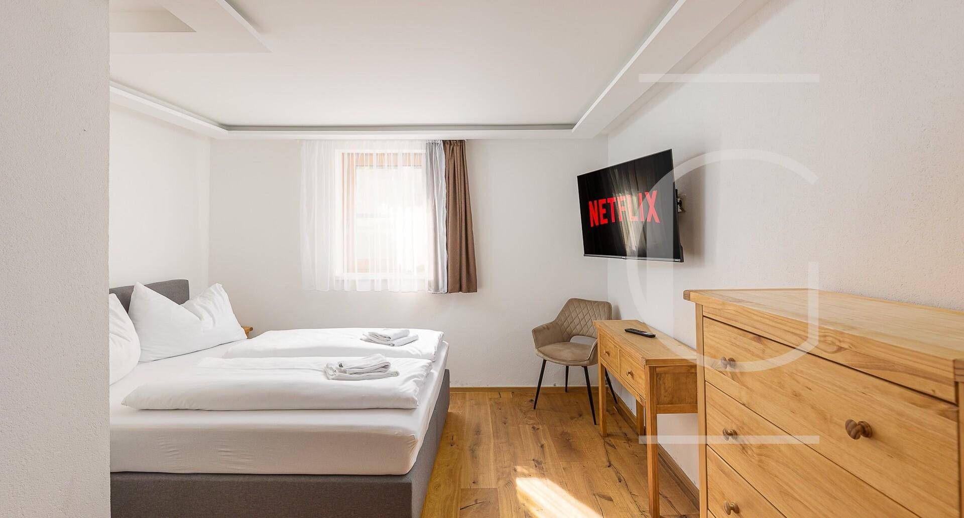 Helles Schlafzimmer mit Doppelbett, Holzboden und einem an der Wand montierten Flachbildfernseher.