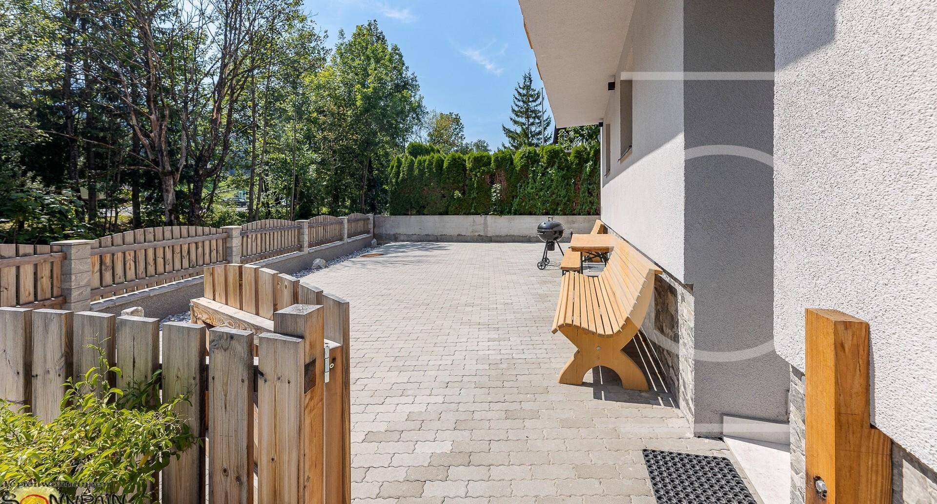 Gemütlicher Außenbereich mit gepflastertem Patio, Holzbank und Grill, umgeben von einer Hecke.