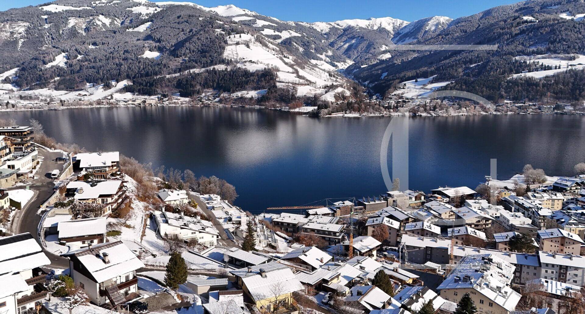 Weitläufiger Blick über eine verschneite Stadt am Seeufer, umgeben von majestätischen Bergen unter blauem Himmel.