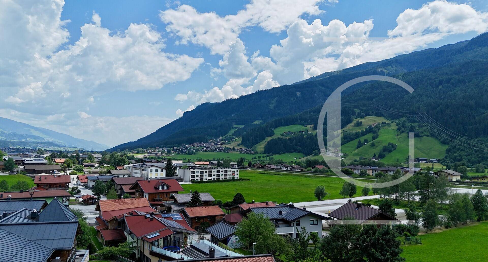 Weitläufiger Panoramablick über ein grünes Tal mit einem Dorf und bewaldeten Bergen unter einem bewölkten Himmel.