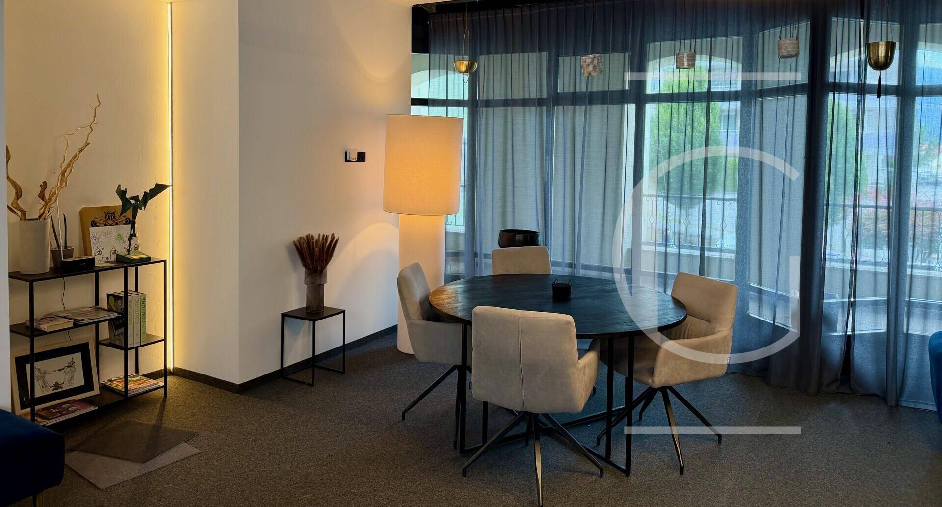 Gemütlicher Loungebereich mit rundem Tisch, bequemen Stühlen und großen Fenstern für entspannte Atmosphäre.
