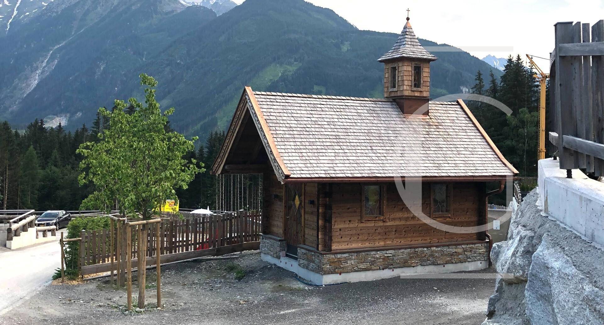Kleine Holzkapelle mit Schindeldach in einer bergigen Landschaft.