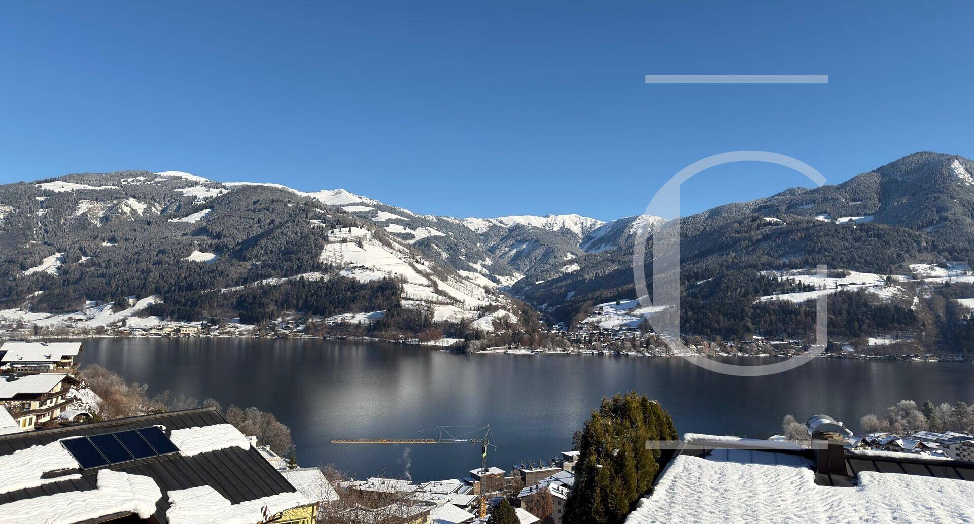 Klarer Blick auf den See und die schneebedeckten Berge, mit Solarpaneelen auf einem Hausdach im Vordergrund.