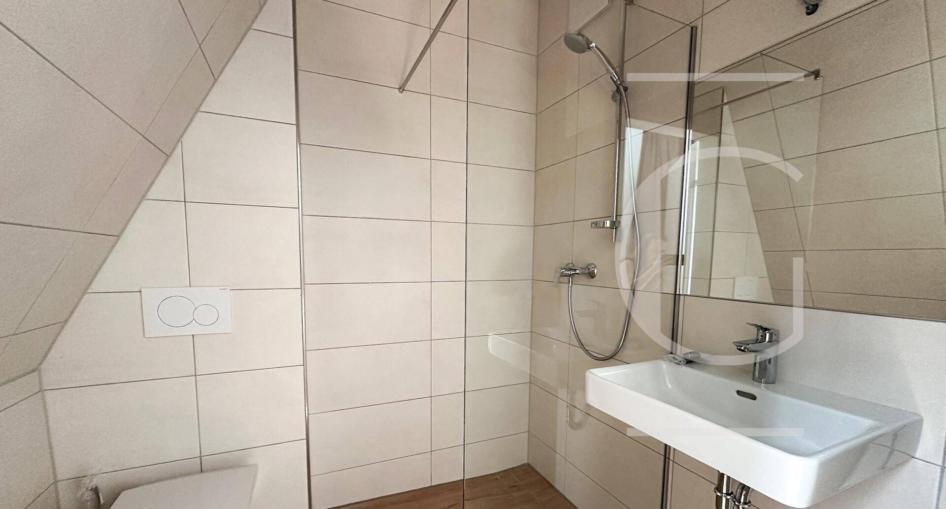 Modernes Badezimmer mit Duschkabine, Waschbecken und hellen Fliesen unter einer Dachschräge.