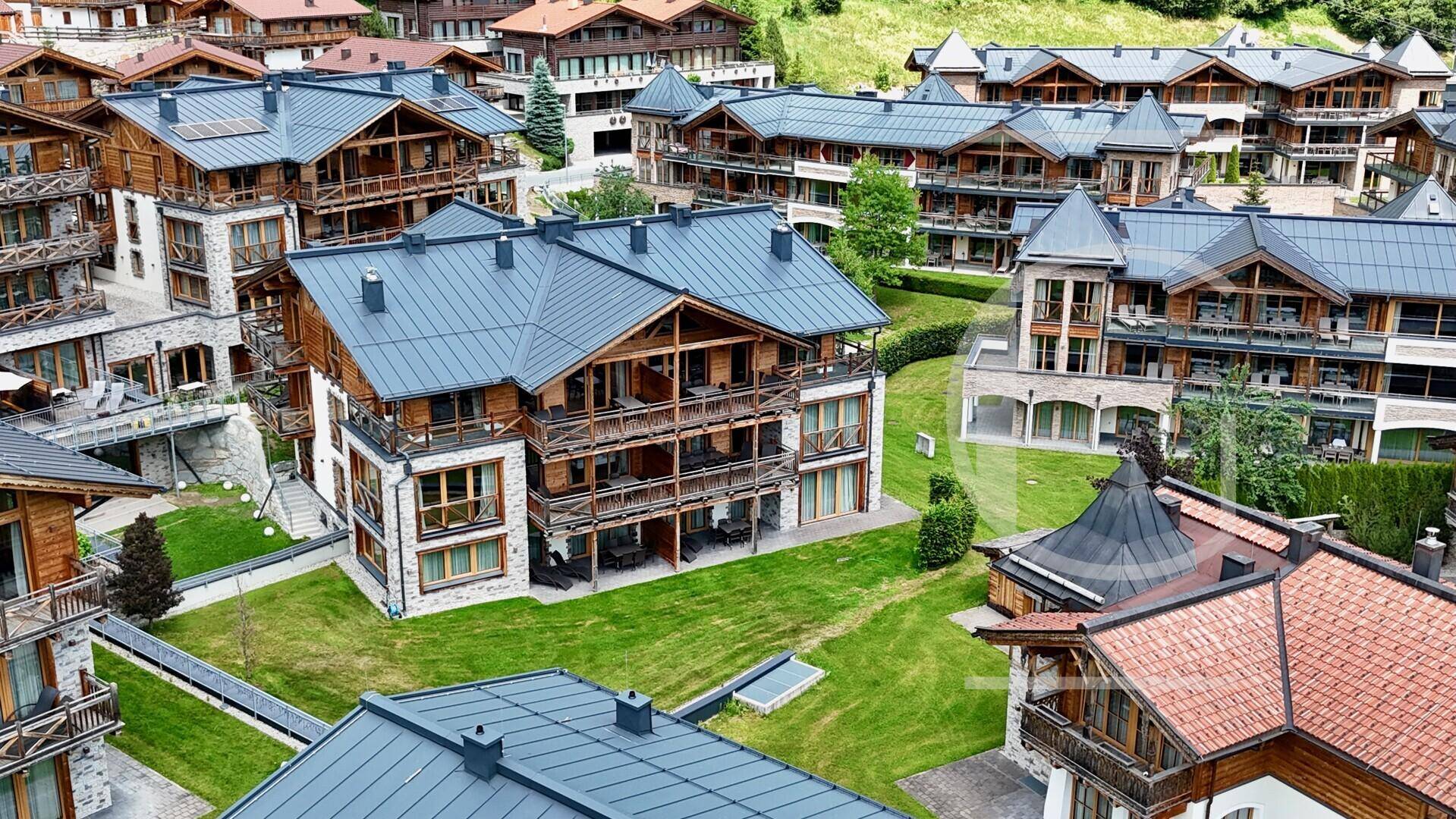 Idyllische Luftaufnahme eines Chalet-Dorfes mit Holzhäusern, grünen Gärten und einem beeindruckenden Bergpanorama.