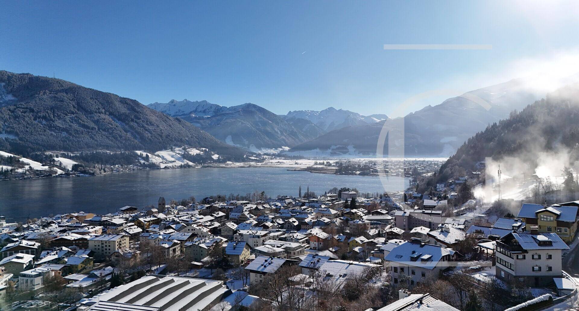 Winterlicher Panoramablick auf eine Stadt am See mit schneebedeckten Dächern und Bergen im Dunst.