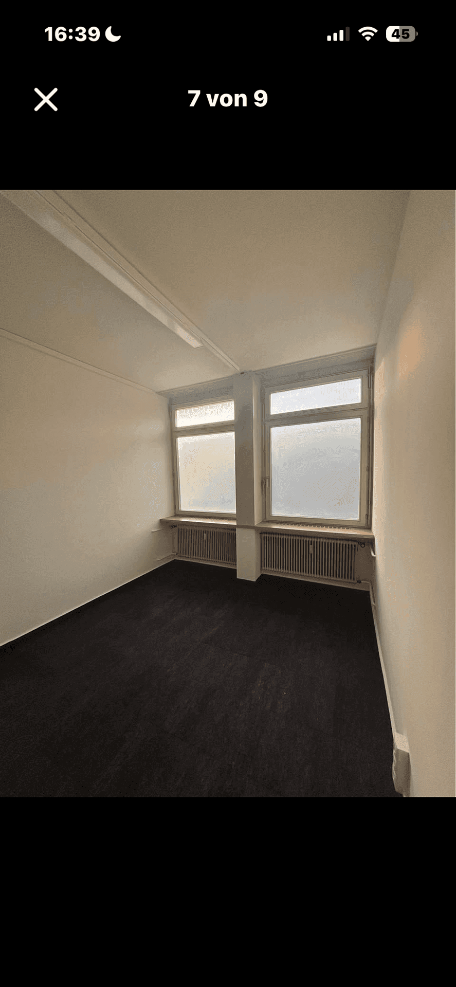 Kleiner Büroraum mit zwei großen Fenstern, Heizkörpern und dunklem Teppichboden.