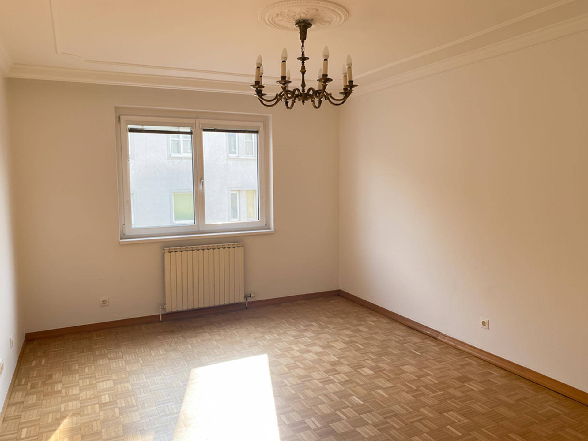 Geräumiges Zimmer mit Parkettboden, Kronleuchter und Heizkörper unter dem Fenster.