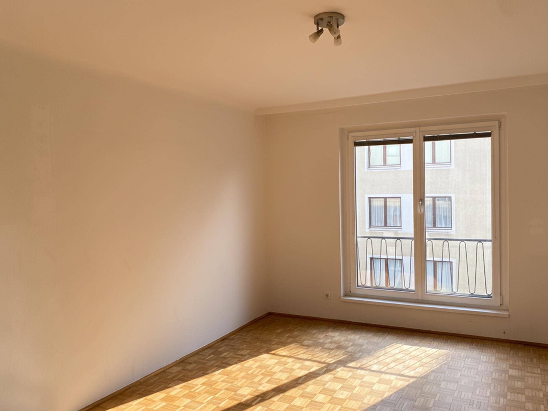 Leeres Zimmer mit Parkettboden und einem Fenster, das Tageslicht hereinlässt.