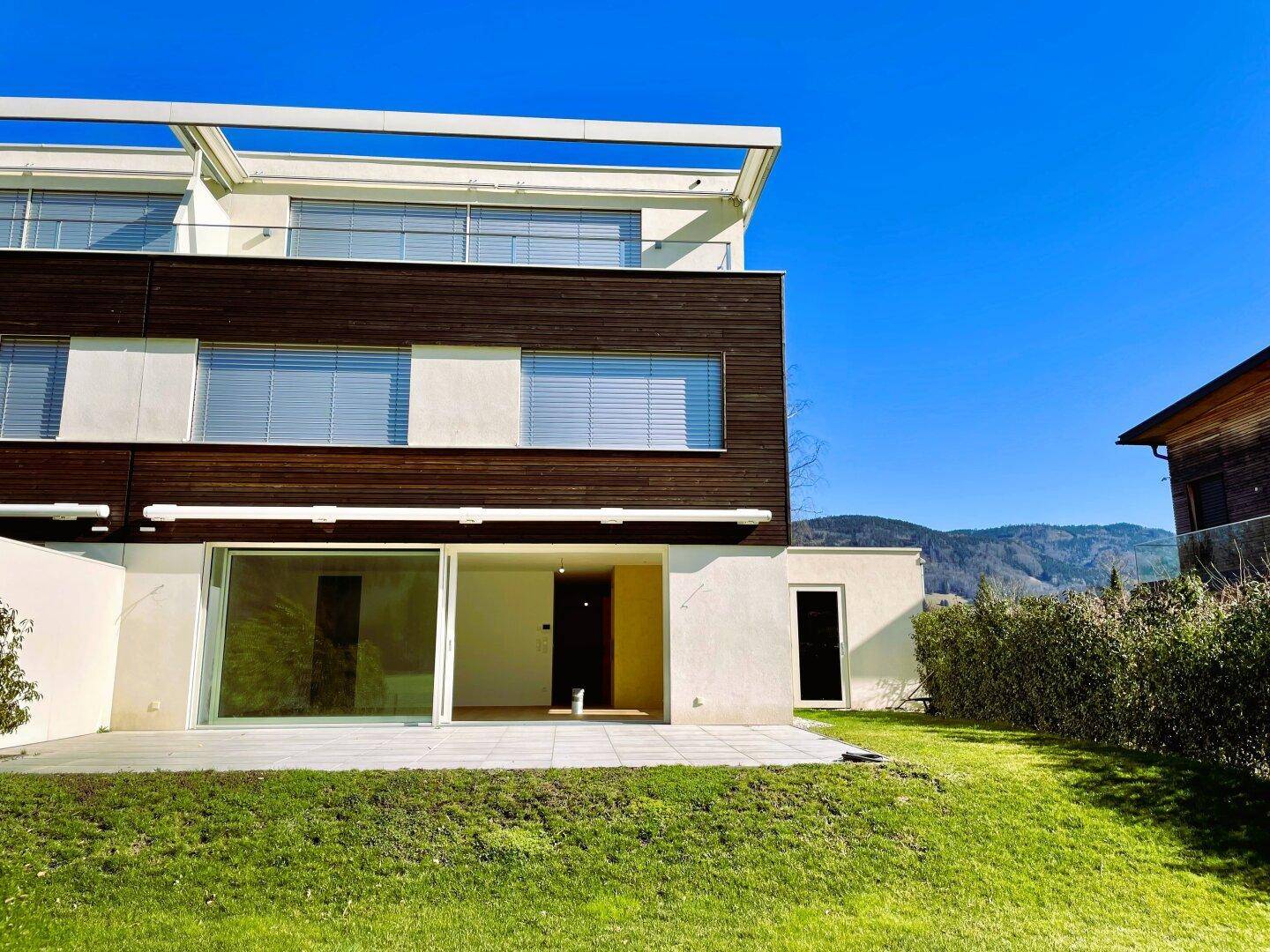 Moderne Architektur eines Neubaus mit großzügiger Terrasse, Rasenfläche und Bergblick in sonniger Lage.