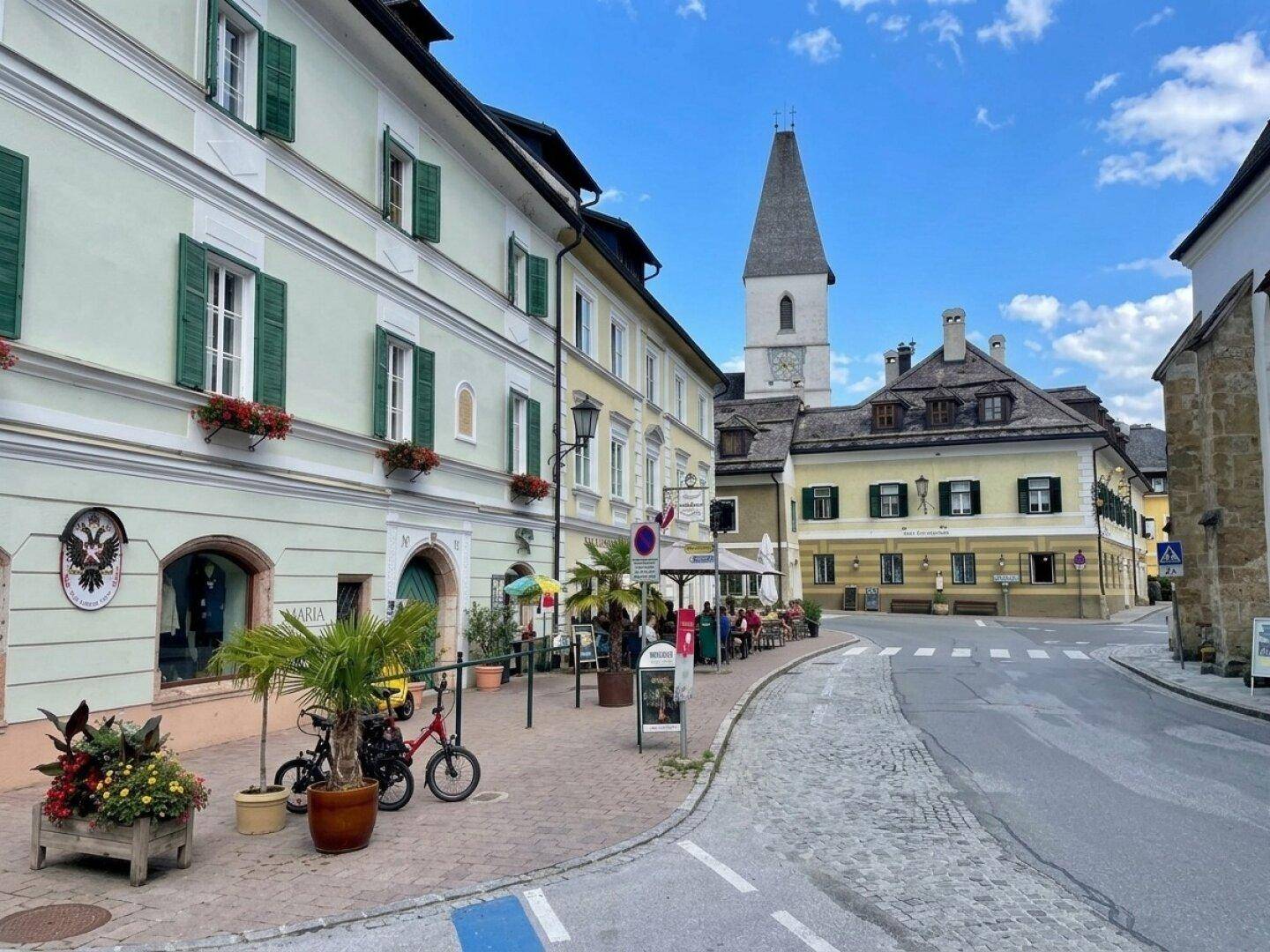 Belebte Gasse mit traditionellen Gebäuden, Geschäften und einem markanten Kirchturm im Hintergrund.