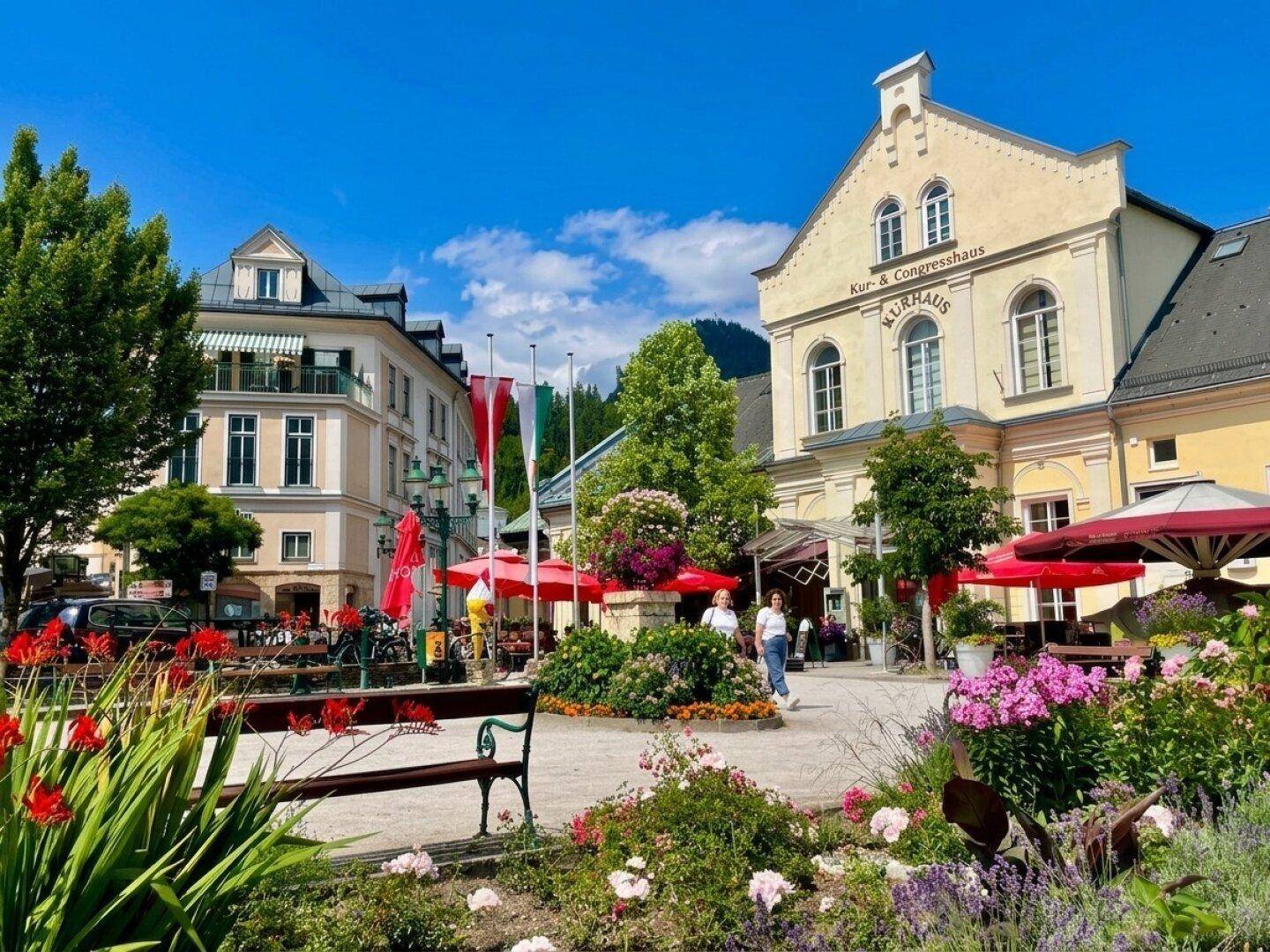 Belebtes Stadtzentrum mit blühenden Blumenbeeten, traditionellen Gebäuden und roten Sonnenschirmen.