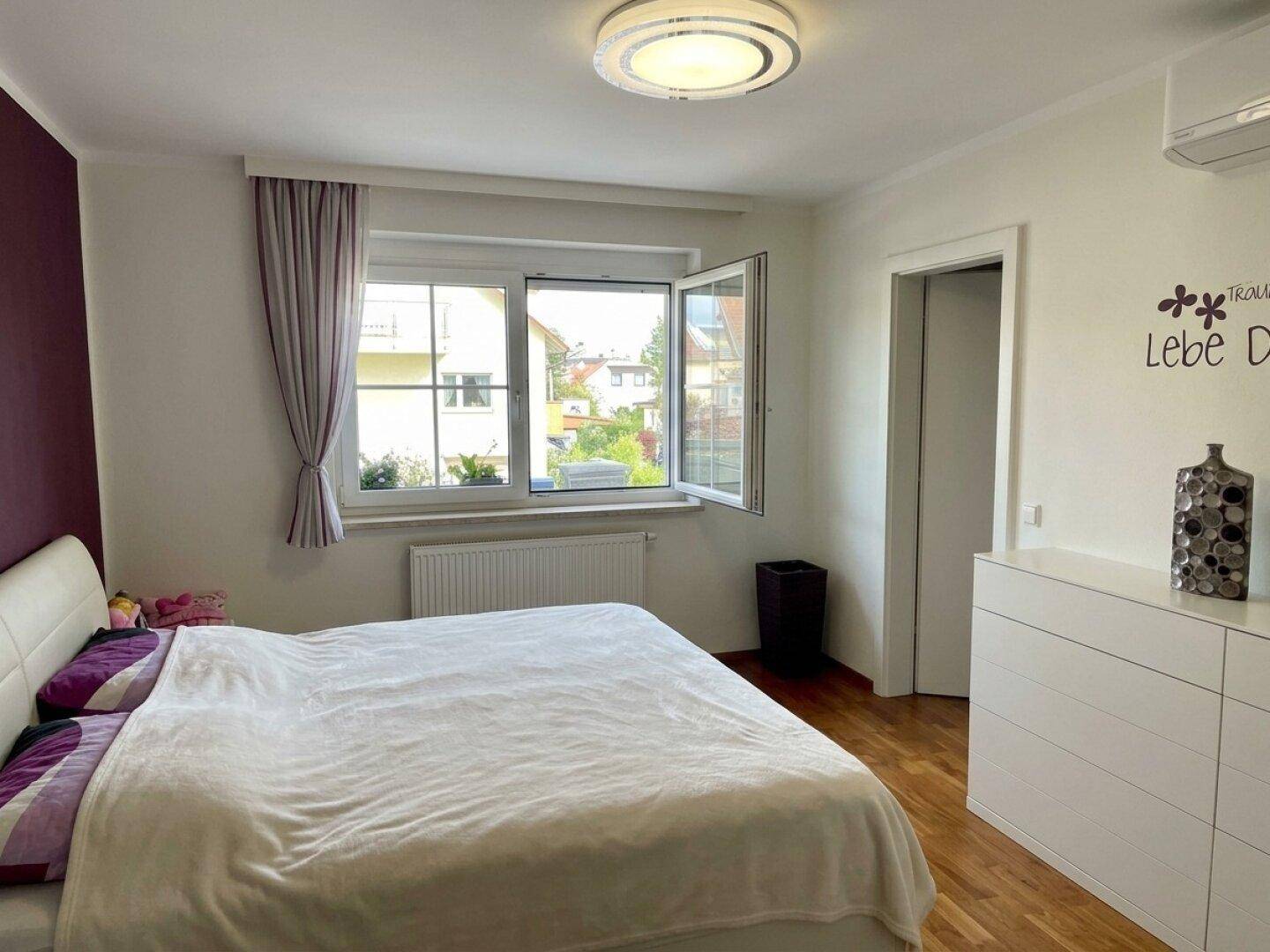 Helles Schlafzimmer mit großem Bett, weißer Kommode und Fenster mit Blick ins Grüne.